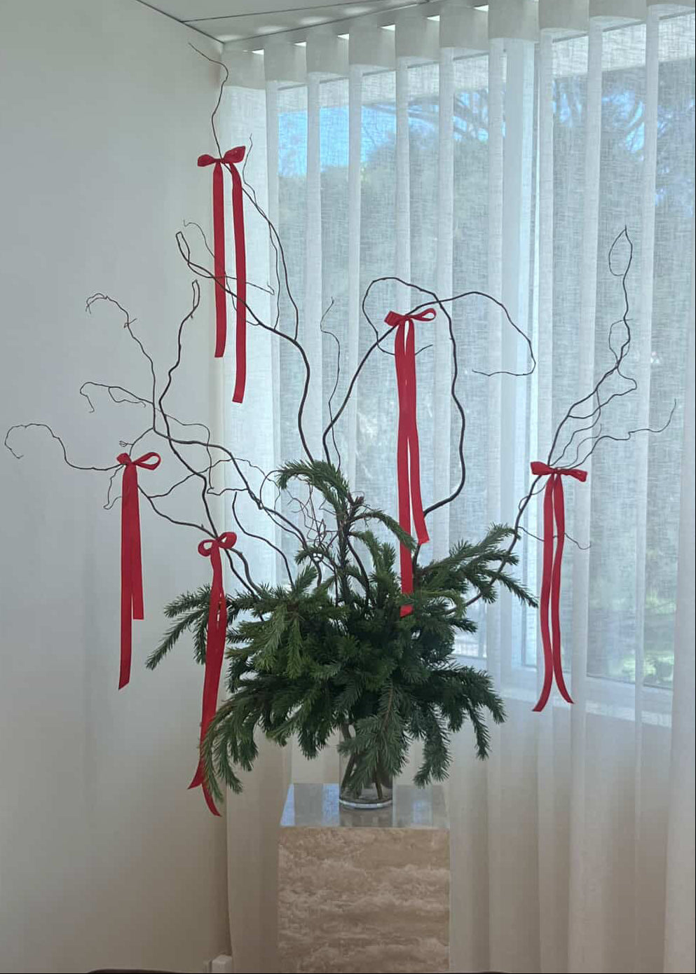 Decorative foliage with red ribbons in a vase against a window with sheer curtains.